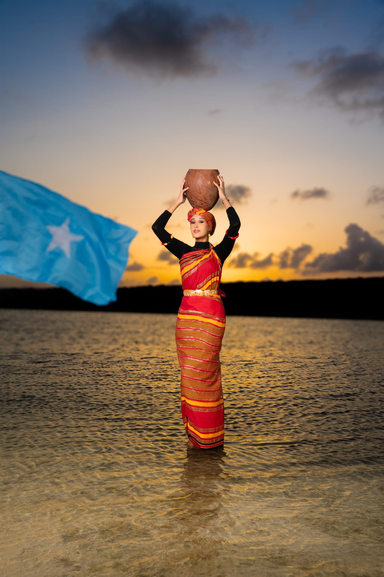 A Somali woman in a red and gold guntiino carries a clay water pot at sunset beside the Somali flag