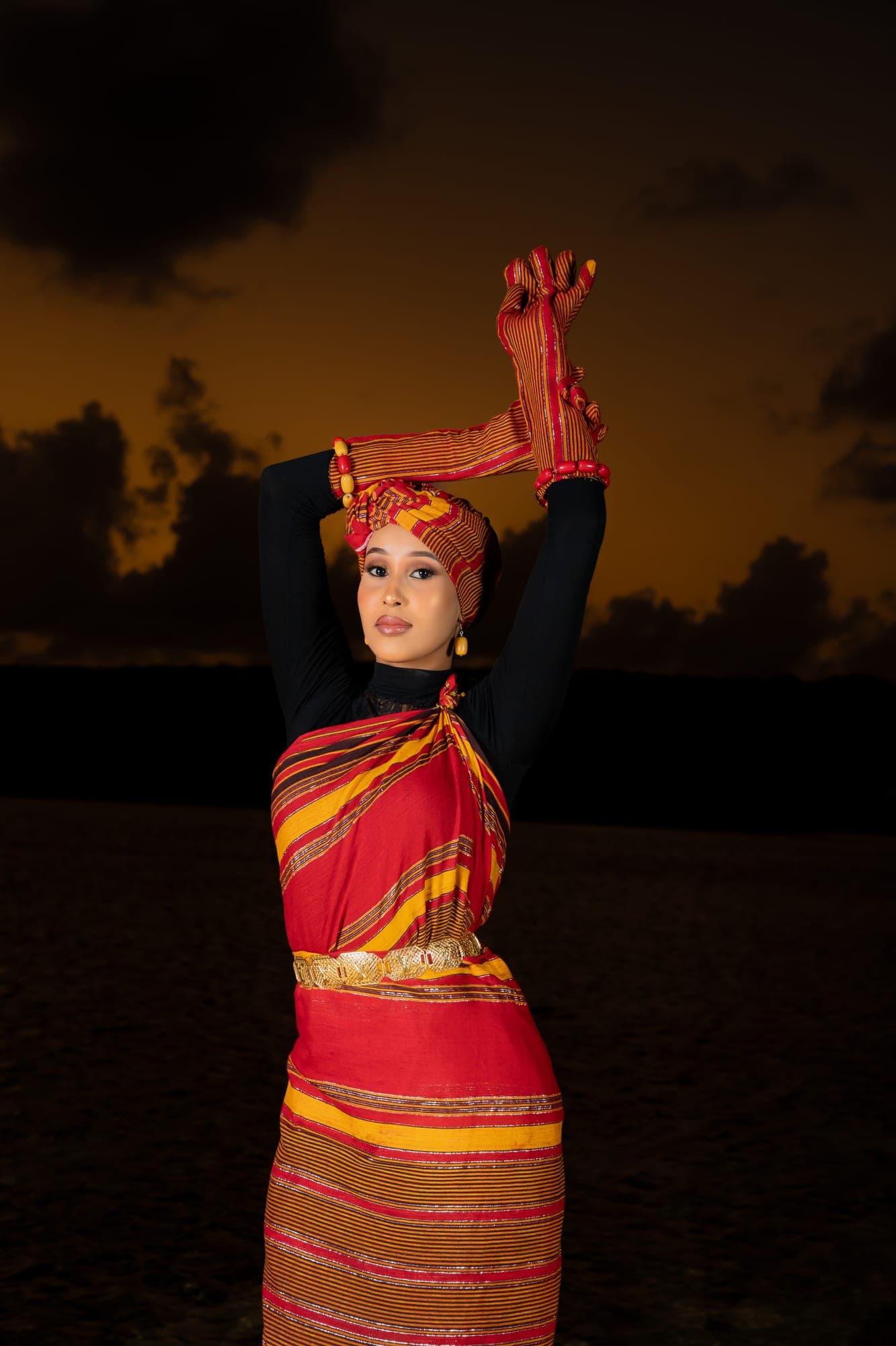 Portrait of a Somali woman with arms raised, wearing a striped guntiino and headwrap at dusk